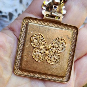 May include: A vintage gold-toned pendant with a square frame and a filigree butterfly design. The pendant has a textured surface and is suspended from a delicate chain. The butterfly is the focal point, with intricate details.