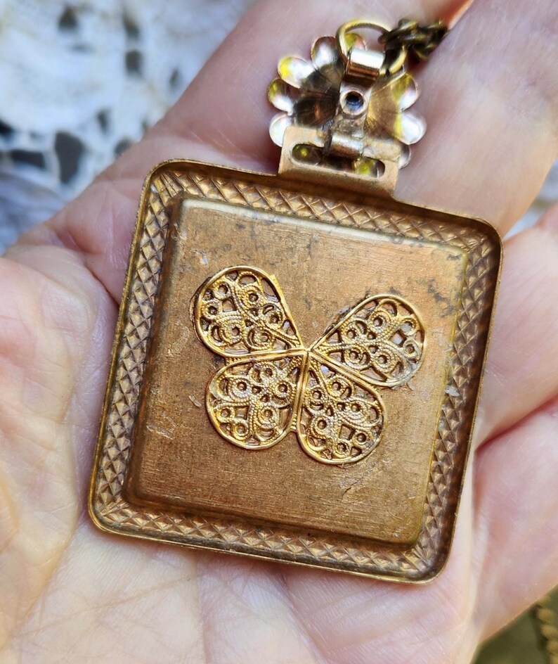May include: A vintage gold-toned pendant with a square frame and a filigree butterfly design. The pendant has a textured surface and is suspended from a delicate chain. The butterfly is the focal point, with intricate details.