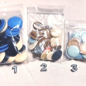 May include: Three small clear plastic bags filled with various decorative buttons. The buttons are in shades of blue, black, cream, and iridescent shell tones. Each bag is labeled with a small, silver, glittery number.