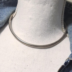 May include: A silver-colored choker necklace with a ribbed design is displayed on a white mannequin. The necklace is circular and sits close to the neck. A denim jacket is visible in the background.