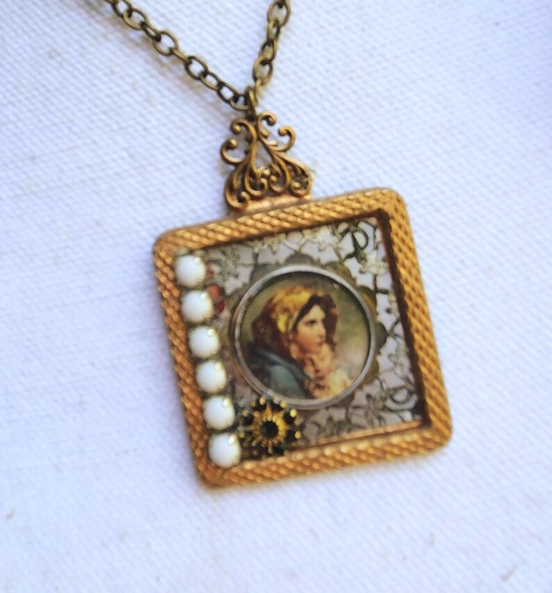 May include: Vintage-style square pendant necklace with a gold-tone frame. The pendant features a portrait of a woman, white beads, and a decorative floral design. The necklace has a delicate chain.