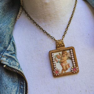 May include: A vintage-style necklace with a rectangular pendant featuring a cherub illustration. The pendant is framed in gold with pink floral accents and a delicate chain. The cherub holds flowers, and the necklace is set against a denim jacket.