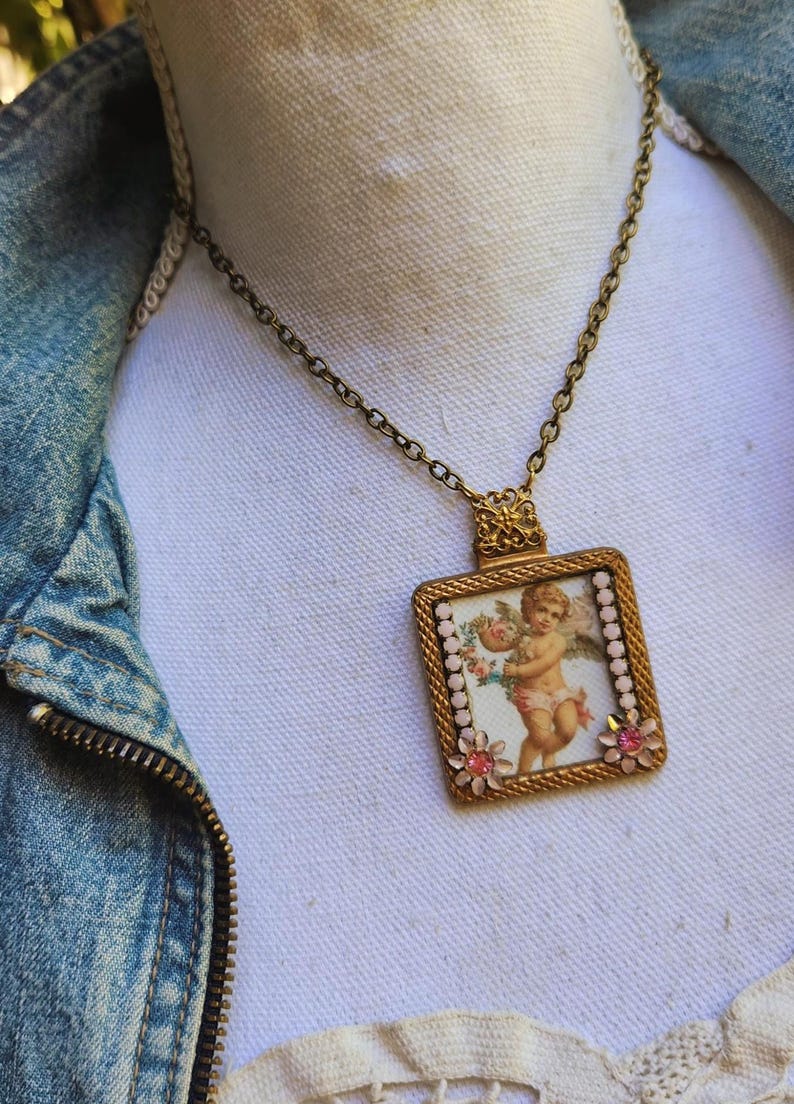 May include: A vintage-style necklace with a rectangular pendant featuring a cherub illustration. The pendant is framed in gold with pink floral accents and a delicate chain. The cherub holds flowers, and the necklace is set against a denim jacket.