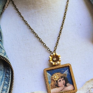 May include: A vintage-style necklace with a gold-toned chain and pendant. The pendant features a small, square frame with a painting of an angel. A gold flower with a small gem is at the top of the pendant.