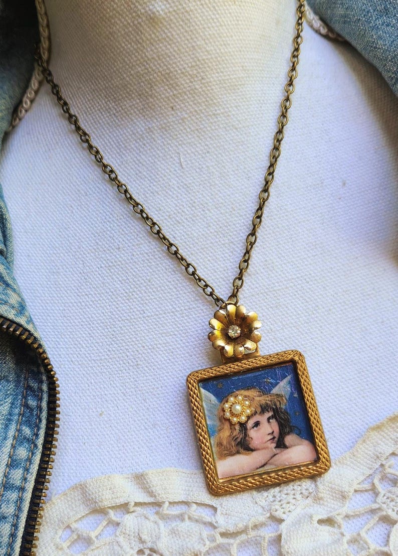 May include: A vintage-style necklace with a gold-toned chain and pendant. The pendant features a small, square frame with a painting of an angel. A gold flower with a small gem is at the top of the pendant.