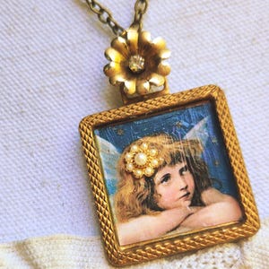 May include: Vintage gold-toned pendant necklace. The pendant features a square frame with a detailed image of a cherub with wings. A cluster of pearls adorns the cherub's hair. The necklace has a gold flower with a crystal center.