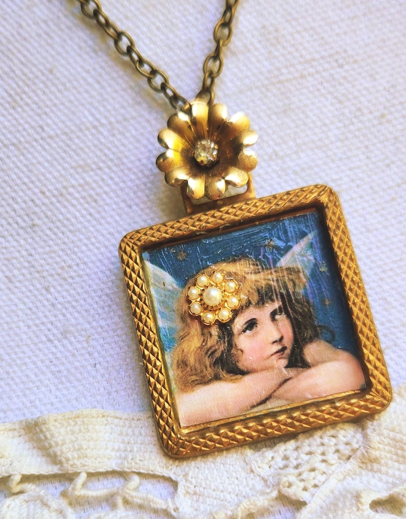 May include: Vintage gold-toned pendant necklace. The pendant features a square frame with a detailed image of a cherub with wings. A cluster of pearls adorns the cherub's hair. The necklace has a gold flower with a crystal center.