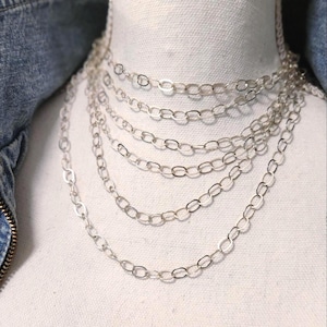 May include: A multi-strand silver chain necklace with oval links is displayed on a white mannequin. The necklace is layered, with each strand of varying lengths. A denim jacket is visible in the background.