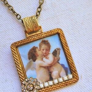 May include: A vintage-style pendant necklace featuring a square, gold-toned frame with a detailed cherub image. The frame is adorned with a crystal flower and white accents. The necklace has a delicate chain.