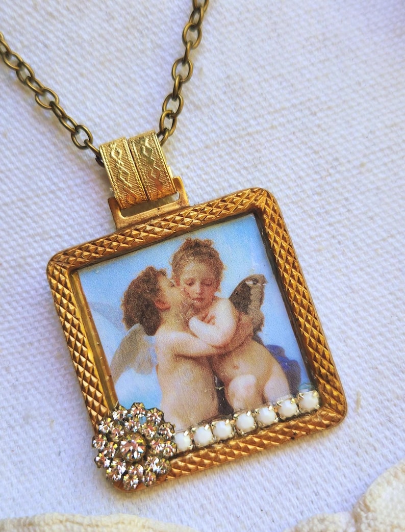 May include: A vintage-style pendant necklace featuring a square, gold-toned frame with a detailed cherub image. The frame is adorned with a crystal flower and white accents. The necklace has a delicate chain.