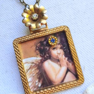 May include: A vintage-style pendant necklace featuring a square pendant with a cherub image. The pendant has a gold-colored frame with a small blue gem. The necklace chain is a dark gold color, and the top of the pendant has a gold flower.