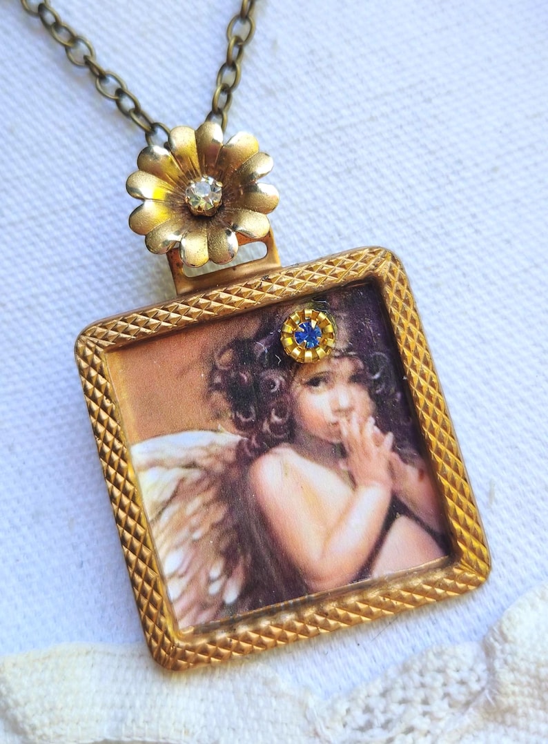 May include: A vintage-style pendant necklace featuring a square pendant with a cherub image. The pendant has a gold-colored frame with a small blue gem. The necklace chain is a dark gold color, and the top of the pendant has a gold flower.