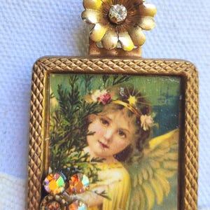 May include: A vintage-style pendant necklace with a square, gold-toned frame. The frame holds a picture of an angel with wings, a floral crown, and a yellow dress. The pendant is adorned with iridescent rhinestones and a gold flower with a central crystal.