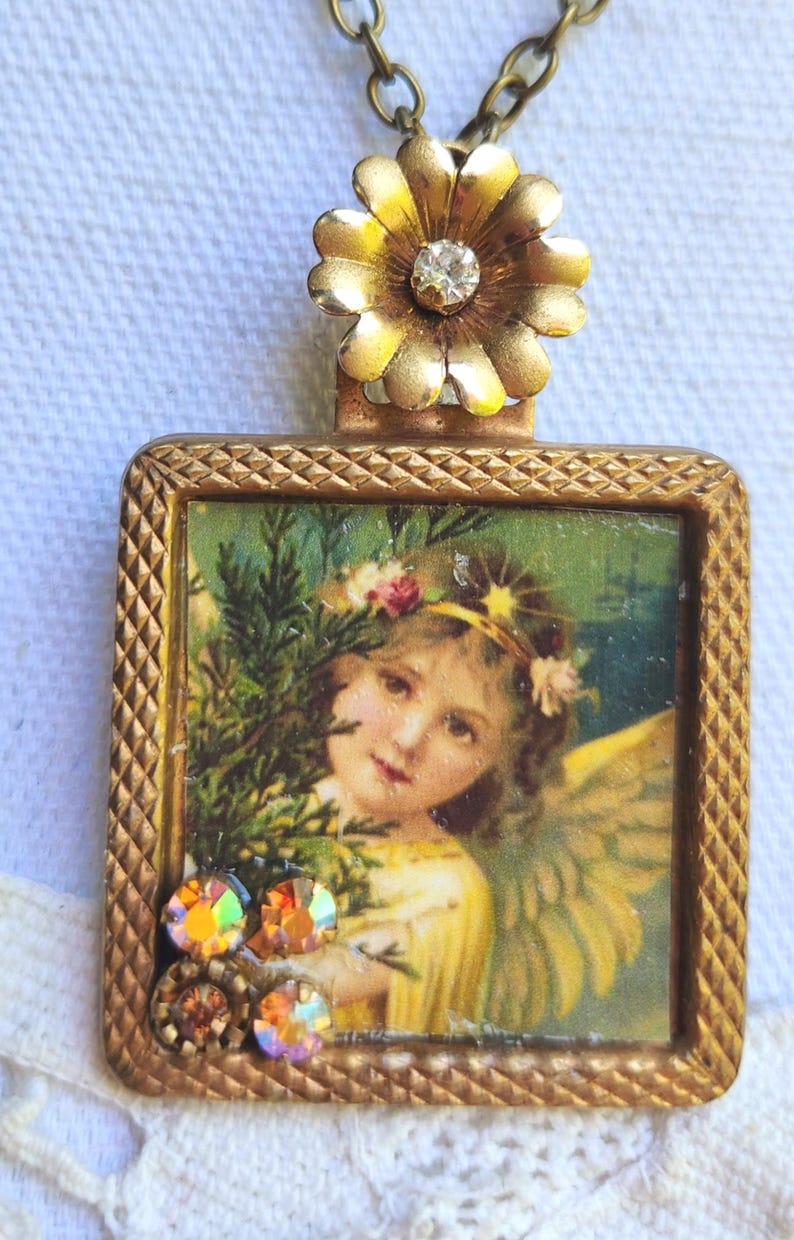 May include: A vintage-style pendant necklace with a square, gold-toned frame. The frame holds a picture of an angel with wings, a floral crown, and a yellow dress. The pendant is adorned with iridescent rhinestones and a gold flower with a central crystal.