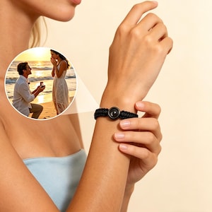 Puede incluir: Una pulsera trenzada negra con una pieza central circular de color oscuro se lleva en la muñeca. La pulsera es el punto focal, con una pequeña imagen insertada de una propuesta en una playa en el fondo.