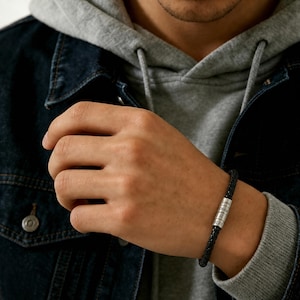 Puede incluir: Una pulsera de cuero trenzado negro con una cuenta cilíndrica de metal plateado. La cuenta está grabada con los nombres Liam y Olivia, un símbolo del zodiaco y la fecha 02.03, aportando un toque personalizado al accesorio.