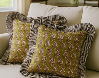 May include: Two decorative throw pillows on a cream-colored sofa. The pillows have a green background with a repeating floral pattern in shades of purple and brown. The pillows are trimmed with a striped ruffle.