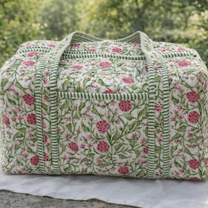 Può includere: Un borsone trapuntato con un motivo floreale di fiori rosa e foglie verdi su sfondo bianco. La borsa ha finiture e manici a righe verdi. La borsa è appoggiata su un panno bianco.