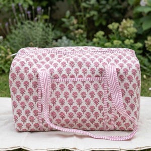 Può includere: Borsa da viaggio rettangolare con motivo floreale rosa e bianco, con manici abbinati e una tasca frontale. La borsa è appoggiata su un panno bianco, con uno sfondo verde di erba e fogliame.
