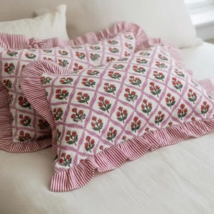 May include: Two decorative throw pillows with a white background and a floral pattern in red and green. The pillows have a ruffled edge with red and white stripes. The pillows are on a white bed.
