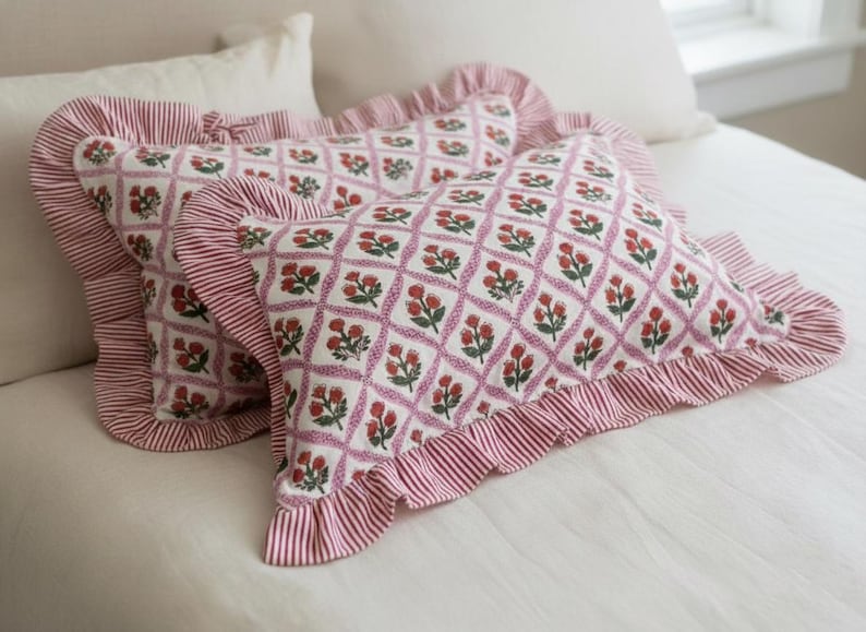 May include: Two decorative throw pillows with a white background and a floral pattern in red and green. The pillows have a ruffled edge with red and white stripes. The pillows are on a white bed.