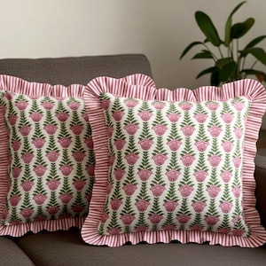 Könnte beinhalten: Zwei quadratische Zierkissen mit floralem Muster. Die Kissen haben einen weißen Hintergrund mit einem sich wiederholenden Muster aus rosa Blüten und grünen Blättern. Die Kanten sind mit einer rosa-weiß gestreiften Rüsche verziert.