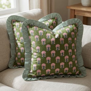 May include: Two square scatter cushions featuring an olive green base with a repeating pattern of pink floral motifs. Each cushion is finished with a ruffled border in a dark green and white striped fabric, creating a decorative edge detail.