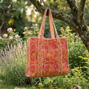 May include: A large tote bag with a floral pattern in shades of orange and pink hangs from a tree branch. The bag has a rectangular shape with a striped handle and trim. The background features a garden with flowers and greenery.