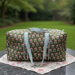 May include: A rectangular quilted duffel bag in olive green featuring a repeating pattern of small pink flowers with green stems. The bag has two light blue and white striped fabric handles and a matching striped zipper closure along the top.