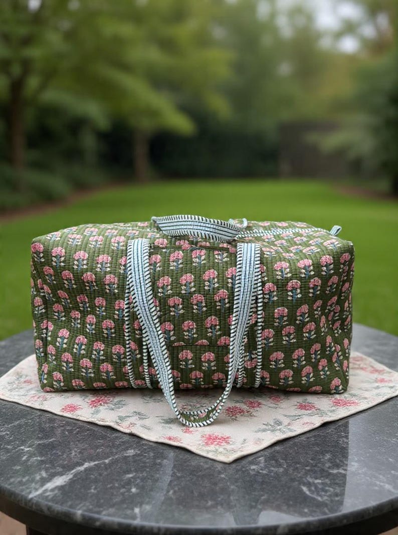 May include: A rectangular quilted duffel bag in olive green featuring a repeating pattern of small pink flowers with green stems. The bag has two light blue and white striped fabric handles and a matching striped zipper closure along the top.