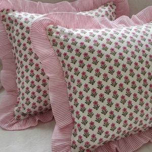 Könnte beinhalten: Zwei dekorative Kissen mit weißem Hintergrund und einem sich wiederholenden Muster aus kleinen rosa Blumen und grünen Blättern. Die Kissen haben rosa und weiß gestreifte Rüschenkanten.