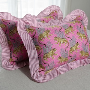 May include: Two decorative throw pillows with a pink background and a pattern of gold leopards. The pillows have a ruffled edge in a pink and white striped fabric. The pillows are on a light gray surface.