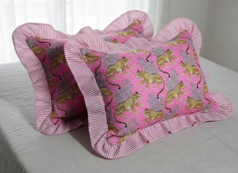 May include: Two decorative throw pillows with a pink background and a pattern of gold leopards. The pillows have a ruffled edge in a pink and white striped fabric. The pillows are on a light gray surface.