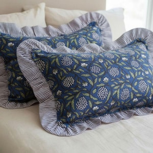 May include: Two decorative rectangular pillows with blue floral patterns and ruffled edges. The pillows have a blue and white striped border. The pillows are on a white bed.
