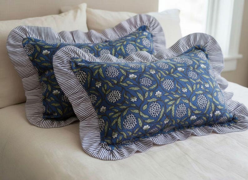 May include: Two decorative rectangular pillows with blue floral patterns and ruffled edges. The pillows have a blue and white striped border. The pillows are on a white bed.