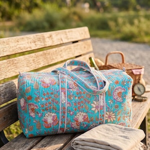 May include: A rectangular quilted duffel bag in a vibrant turquoise fabric featuring a repeating floral pattern in shades of orange, pink, and white. The bag includes two matching fabric handles and a striped trim along the seams and edges.