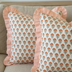 May include: Two decorative throw pillows with a floral pattern. The pillows have a white background with a repeating pattern of orange flowers and green leaves. The edges are trimmed with a ruffled border in a striped orange and white fabric.