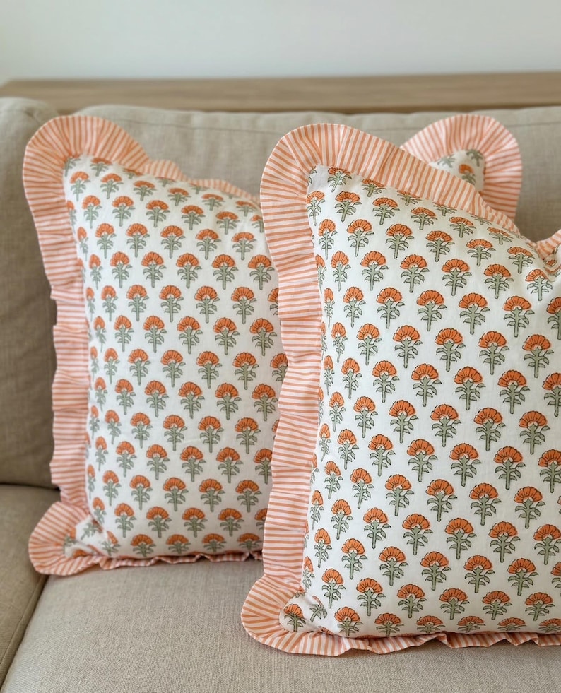 May include: Two decorative throw pillows with a floral pattern. The pillows have a white background with a repeating pattern of orange flowers and green leaves. The edges are trimmed with a ruffled border in a striped orange and white fabric.
