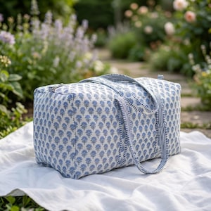 May include: A rectangular duffel bag featuring a repeating blue floral block print pattern on a white background. The bag includes two matching blue and white striped fabric handles and a silver-toned metal zipper closure along the top edge.