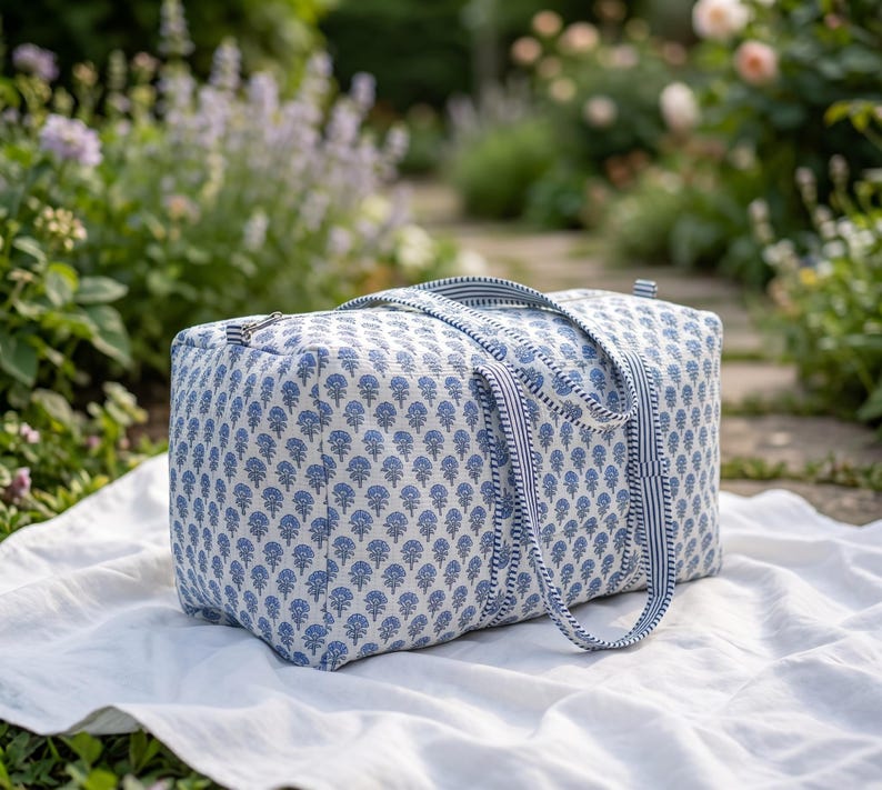 May include: A rectangular duffel bag featuring a repeating blue floral block print pattern on a white background. The bag includes two matching blue and white striped fabric handles and a silver-toned metal zipper closure along the top edge.