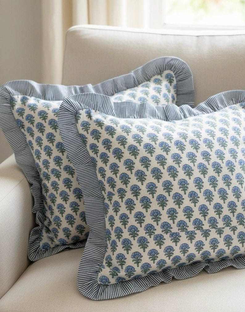 May include: Two decorative throw pillows with a floral pattern in shades of blue and green on a cream background. The pillows have a ruffled edge with blue and white stripes. The pillows are on a cream colored sofa.