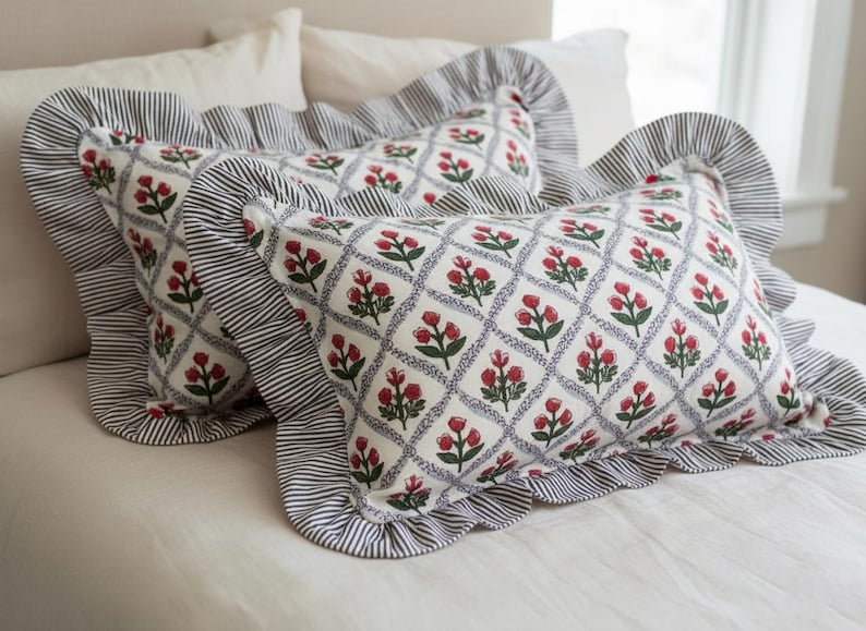 May include: Two decorative throw pillows with a floral pattern of red flowers and green leaves on a white background. The pillows have a blue diamond grid design and are trimmed with a ruffled edge of blue and white stripes.