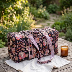 May include: A rectangular quilted duffel bag featuring a black base with a multi-colored floral print in shades of pink, orange, and cream. The bag includes two striped fabric handles and a zipper closure along the top edge.