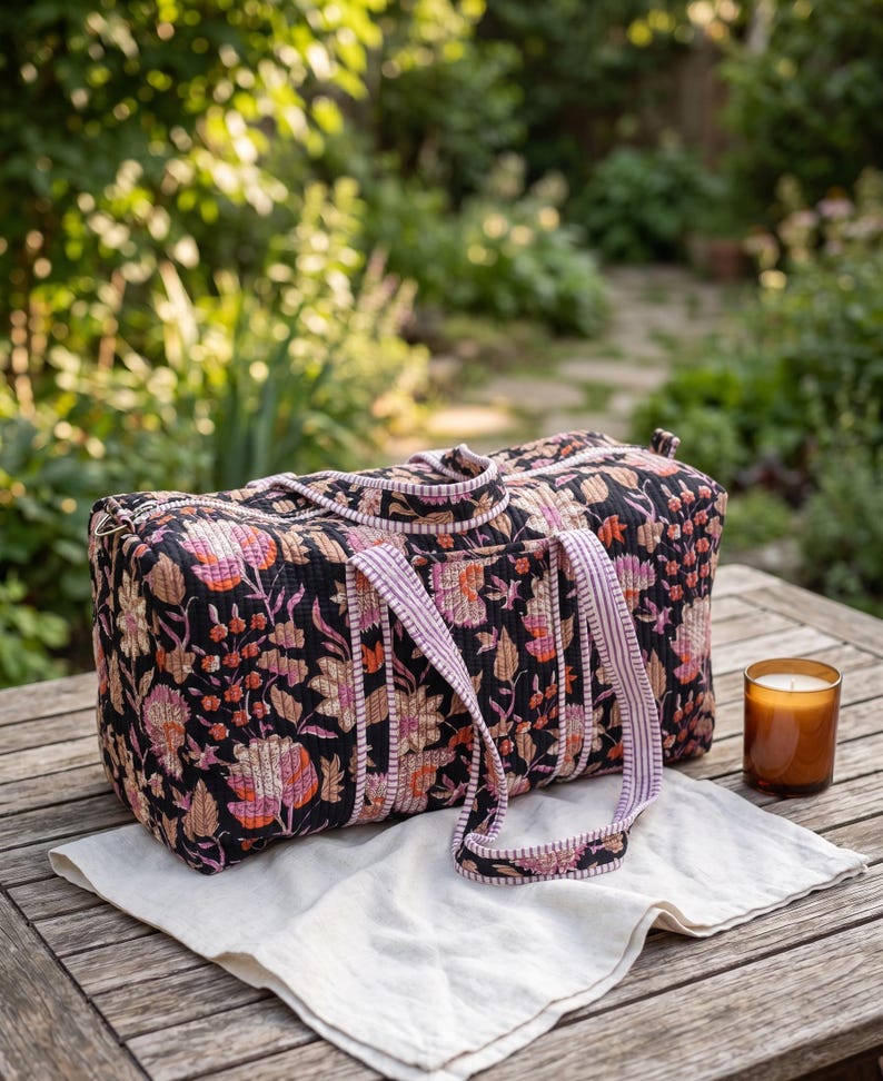 May include: A rectangular quilted duffel bag featuring a black base with a multi-colored floral print in shades of pink, orange, and cream. The bag includes two striped fabric handles and a zipper closure along the top edge.