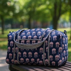 Puede incluir: Una bolsa de lona acolchada azul marino con un estampado floral rosa. La bolsa tiene una asa y ribetes a rayas. La bolsa está sobre un banco de madera en un parque.