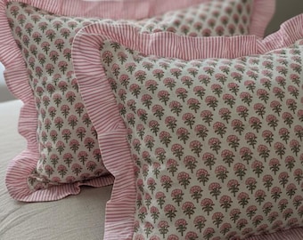 May include: Two decorative pillows with a floral pattern in pink and gray on a cream background. The pillows have a ruffled edge and a striped border in pink and white. The pillows are on a neutral-colored bedspread.