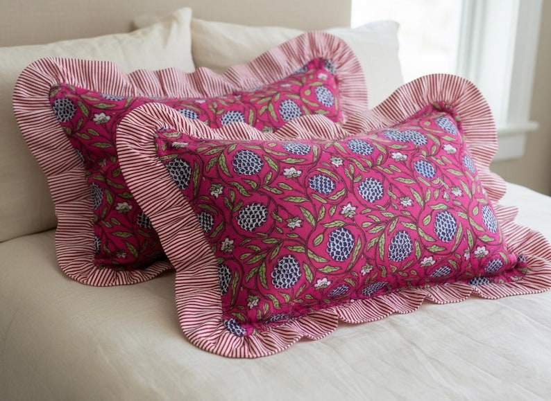 May include: Two decorative throw pillows with a vibrant pink floral pattern and ruffled edges. The pillows feature a repeating design of blue and green floral elements on a pink background, framed by a red and white striped border.