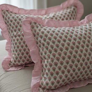 May include: Two decorative pillows with a floral pattern in pink and gray. The pillows have a ruffled edge and a striped border in pink and white. The pillows are on a neutral-colored surface.