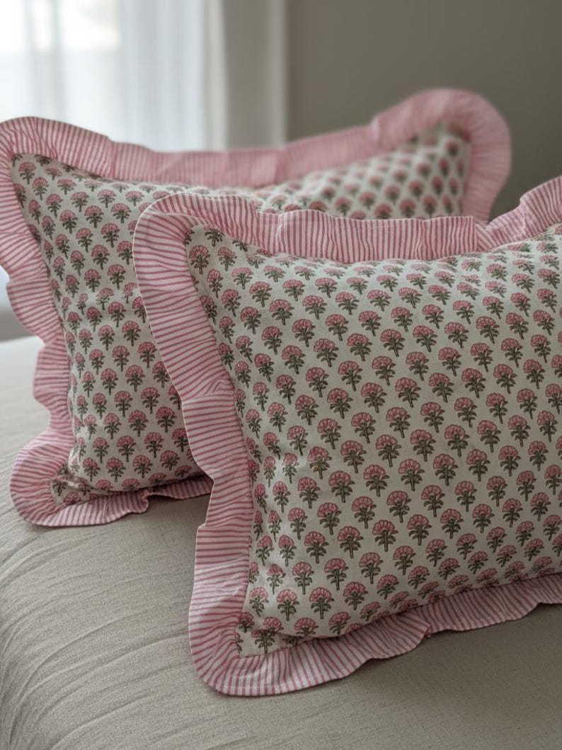May include: Two decorative pillows with a floral pattern in pink and gray. The pillows have a ruffled edge and a striped border in pink and white. The pillows are on a neutral-colored surface.