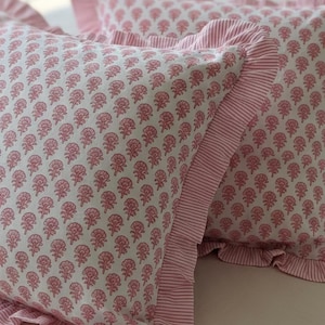 May include: Two decorative pillows with a white background and a repeating pink floral pattern. The pillows have a ruffled edge and a striped border in pink and white. The pillows are on a white surface.